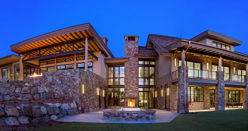 Exterior of a custom luxury home in Wolf Creek Ranch, Utah .near Park City. The view features custom stone masonry, floor to ceiling windows, an outdoor two story chimney with fireplace and hot tub.